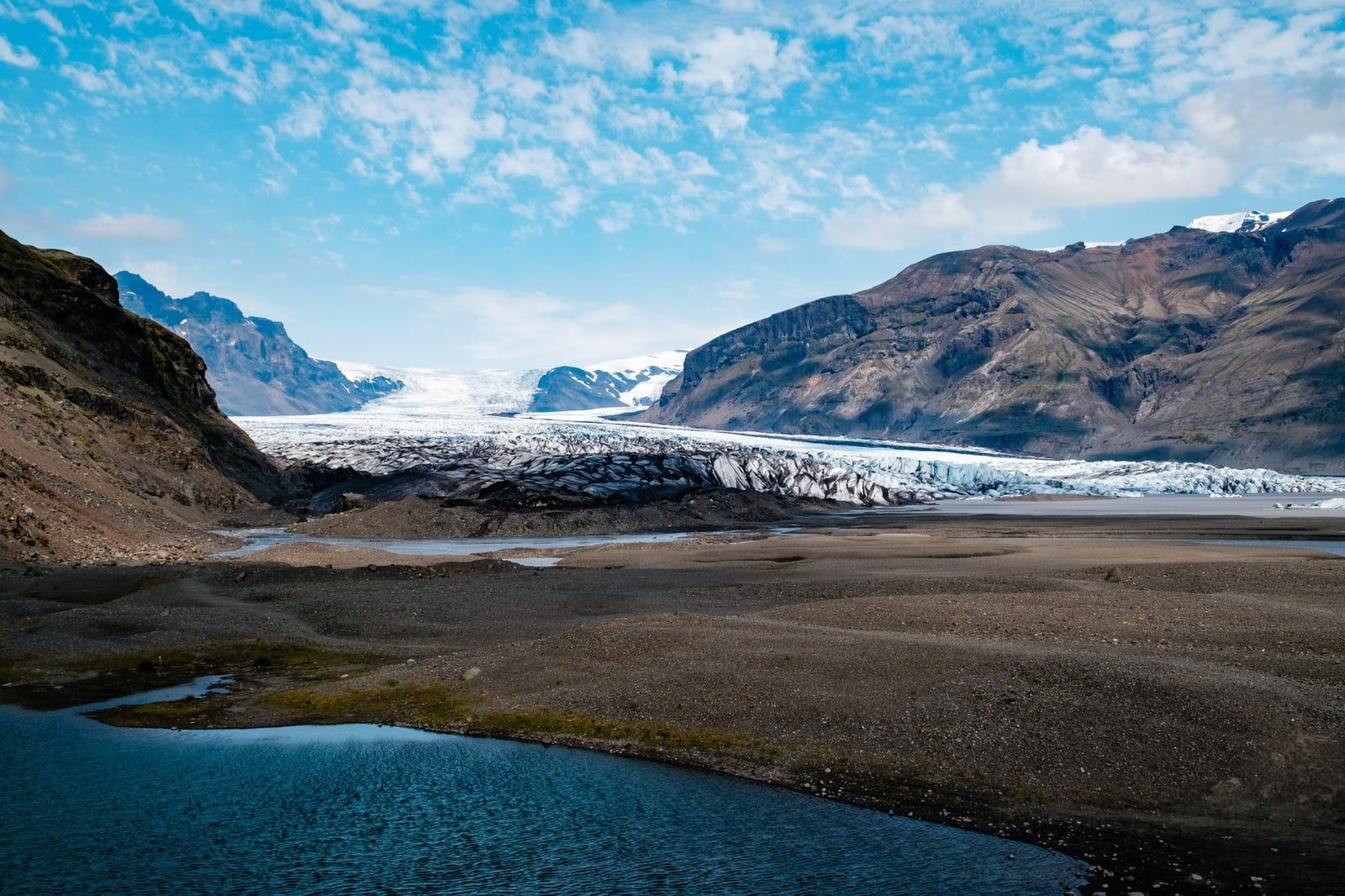Skaftafell Glacier flows through a wide mountain valley toward a proglacial lake under a partly cloudy sky.