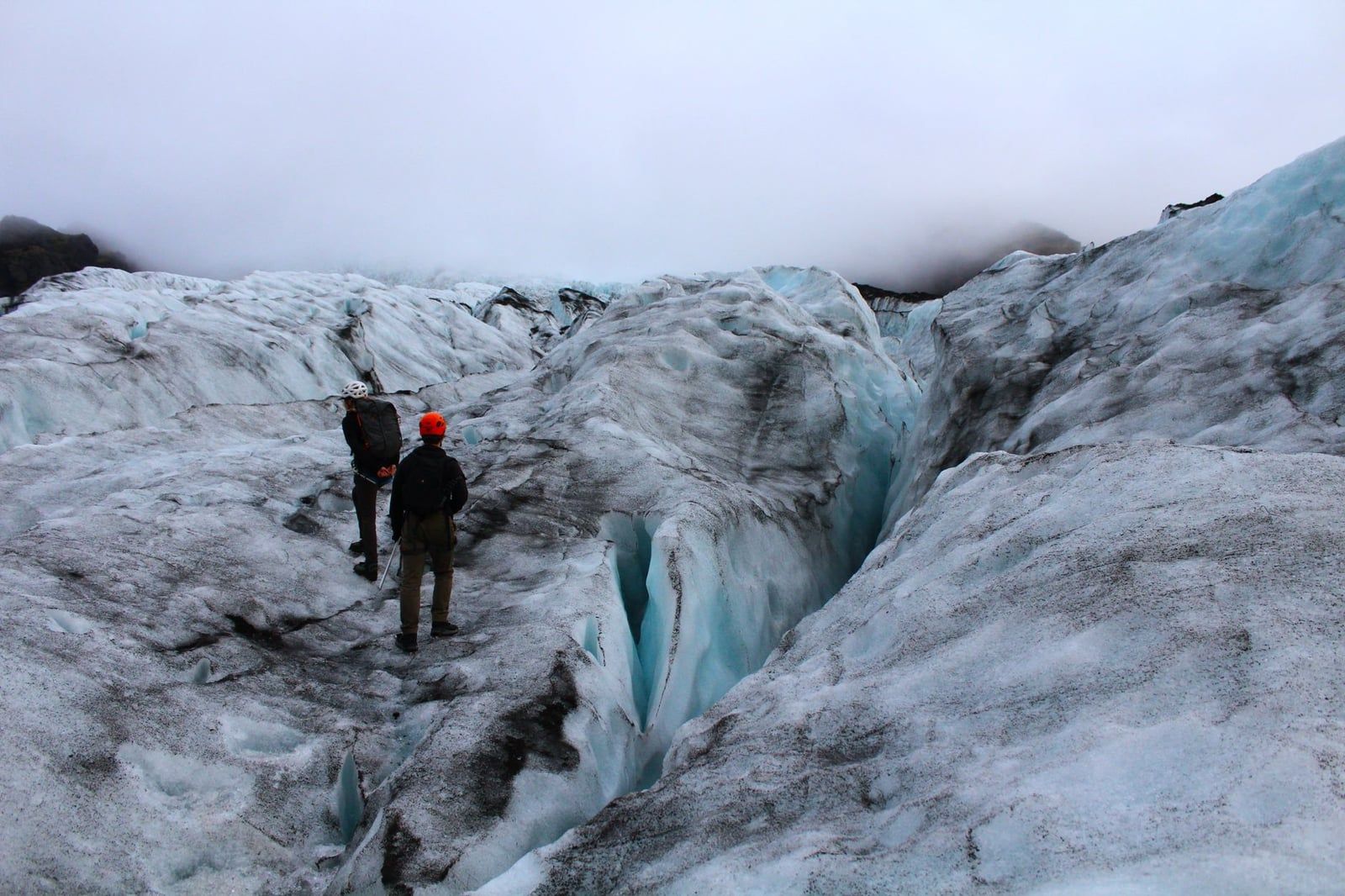 Two hikers wearing safety helmets stand near a deep blue crevasse while traversing a rugged glacier in Iceland.