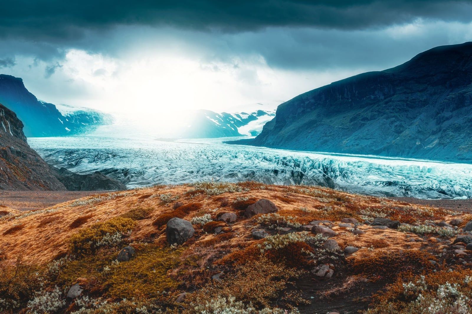 Panoramablick von einem moosbewachsenen Kamm über die gewaltige Eisfläche des Skaftafell-Gletschers, der unter dramatischem Himmel zwischen Bergabhängen fließt.